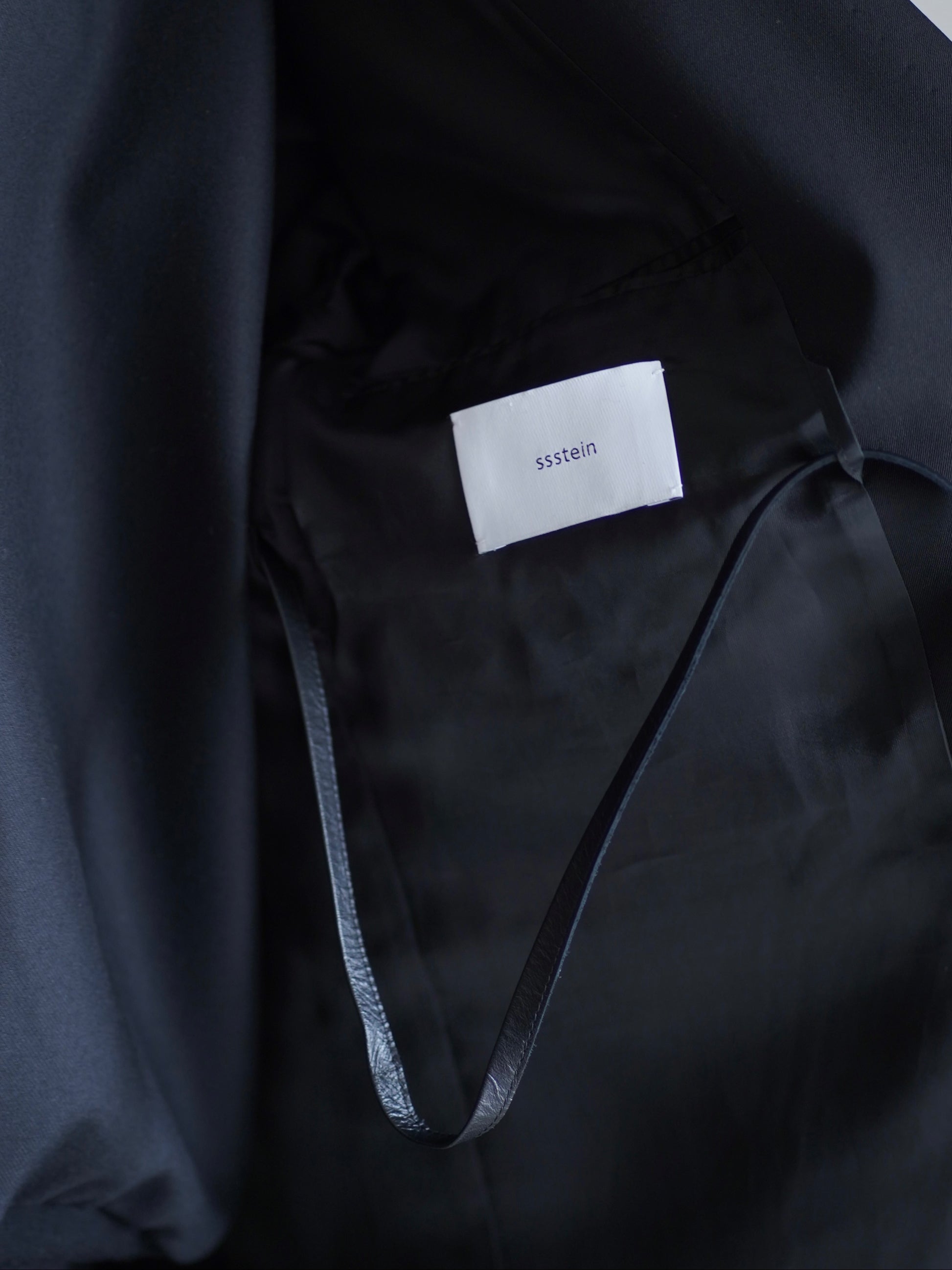 sssteinのOVERSIZED SINGLE BREASTED LONG JACKETのカラーはBLACKの左胸のジャケットの内側のブランドロゴとレザーコードを収納した際の画像です。こちらの商品は通販での購入が可能です。
